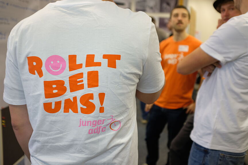 Shirts Junger ADFC Jugendforum ADFC NRW 2025 Ein Mensch mit einem T-Shirt, auf dem hinten der Spruch "Rollt bei uns!" steht.