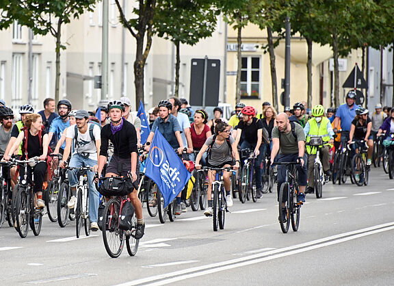 Teilnehmende der ADFC-Fahrradsternfahrt mit blauer ADFC-Fahne fahren gemeinsam auf einer breiten Straße. Menschen jeden Alters mit Helmen und teilweise Warnwesten demonstrieren für bessere Radinfrastruktur. Im Hintergrund säumen Bäume die Straße.