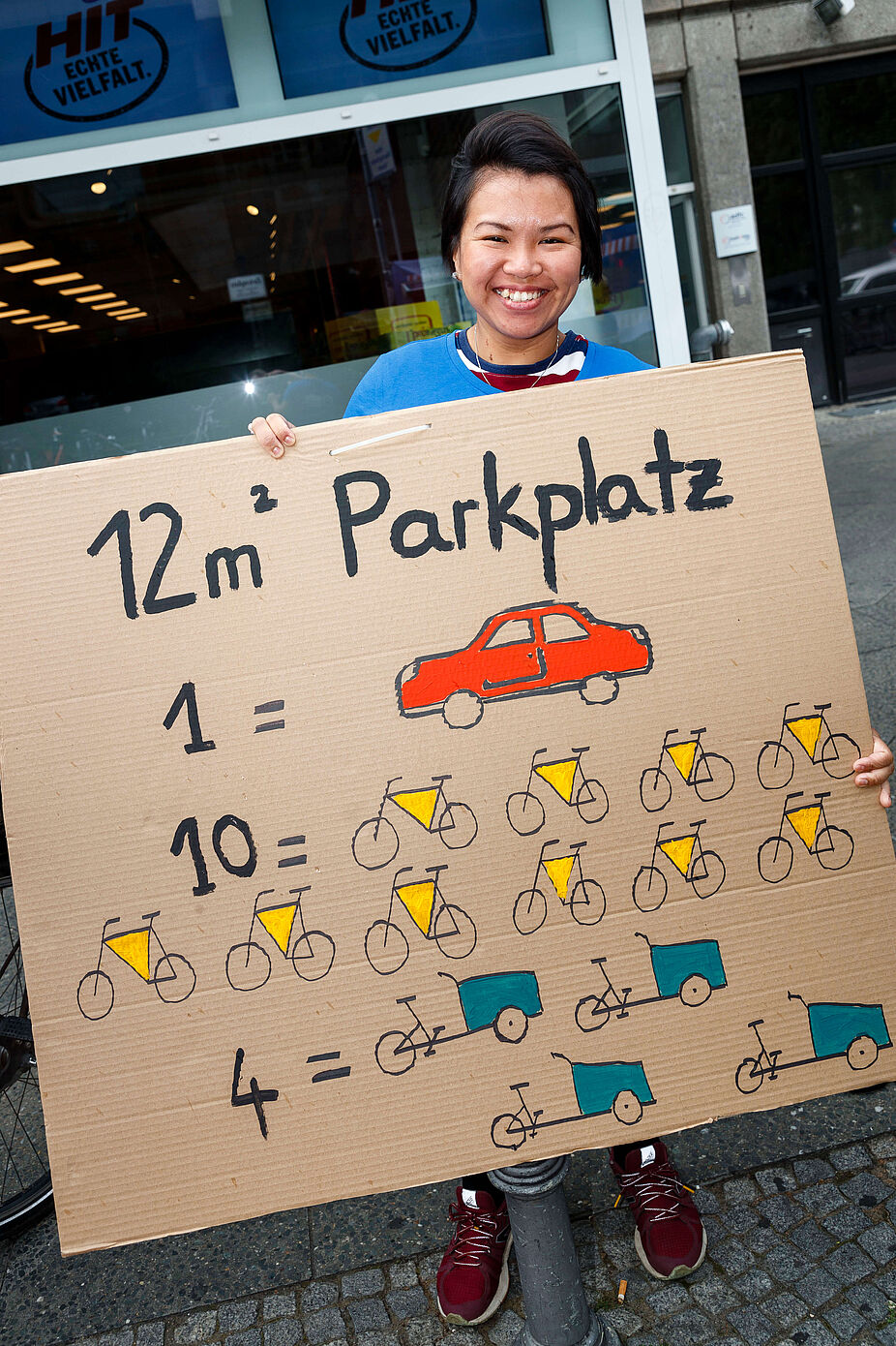 Beim Park(ing) Day werden Parkplätze umgewidmet. Frau hält Plakat mit Aufschrift: Ein Autoparkplatz = 12 Fahrräder = 4 Lastenräder